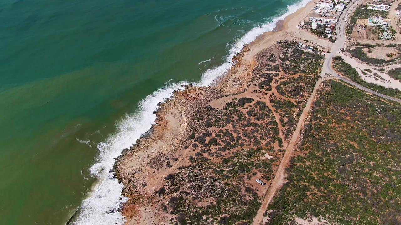 Aerial frame over Buzzards Beach East Cape BCS Mexico with a fresh line of white foam along the beach, green ocean to the left, scrubland to the right, and no visible people.