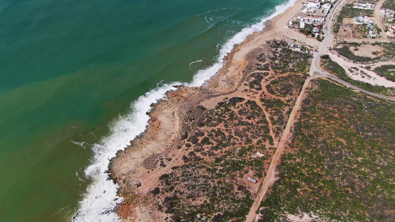 Aerial frame over Buzzards Beach East Cape BCS Mexico showing the wave foam pulled along the shore, patchy coastal vegetation, tan dirt paths, nearby buildings, and no visible people.
