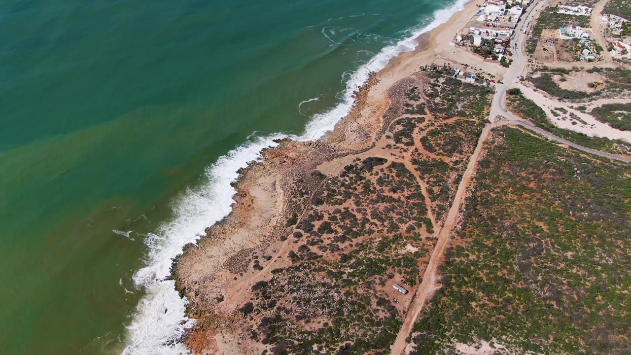 Aerial coastline with white surf, rocky sand, scrub vegetation, and inland dirt road.