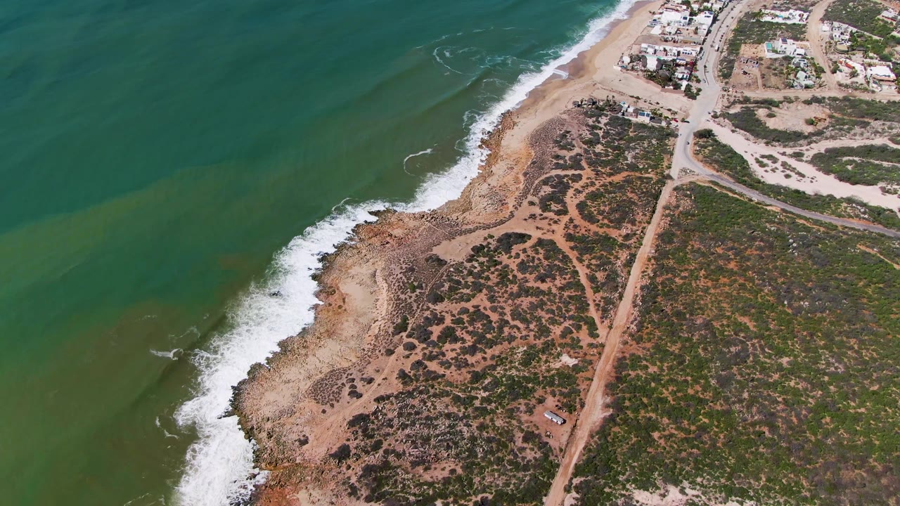 Aerial frame from Buzzards Beach East Cape BCS Mexico showing green water, white foam along the sandy rocky edge, inland scrub, dirt roads, small buildings, and no visible people.