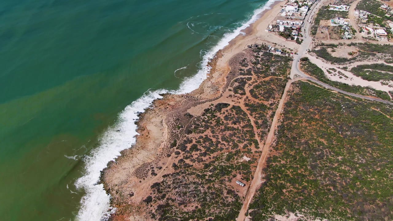 Drone view over a curving beach with white surf, green water, scrub vegetation, and a dirt road inland.
