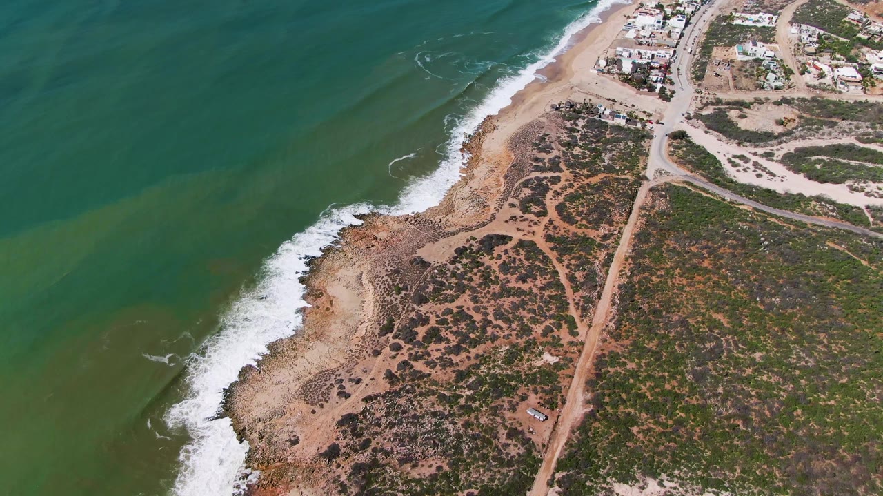 Aerial frame from Buzzards Beach East Cape BCS Mexico showing a slightly later surf pattern against the rocky sand edge, crossed dirt paths, sparse vegetation, buildings, and no visible people.