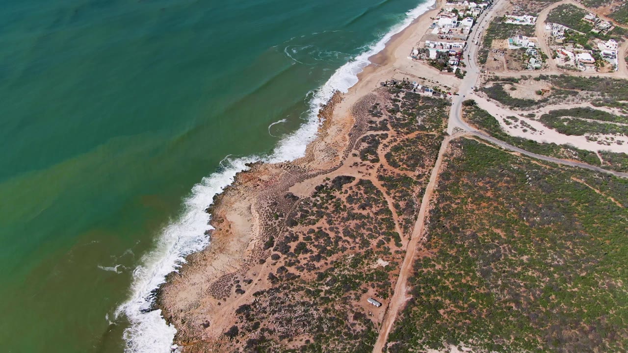 Aerial frame at Buzzards Beach East Cape BCS Mexico showing green water, white surf, rocky sandy shoreline, scrubland, a dirt road, distant buildings, and no visible people.