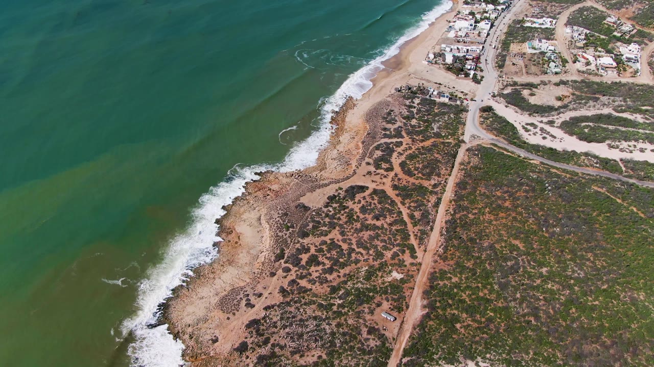 Drone view of surf wrapping around a rocky point with a coastal dirt road and low buildings inland.