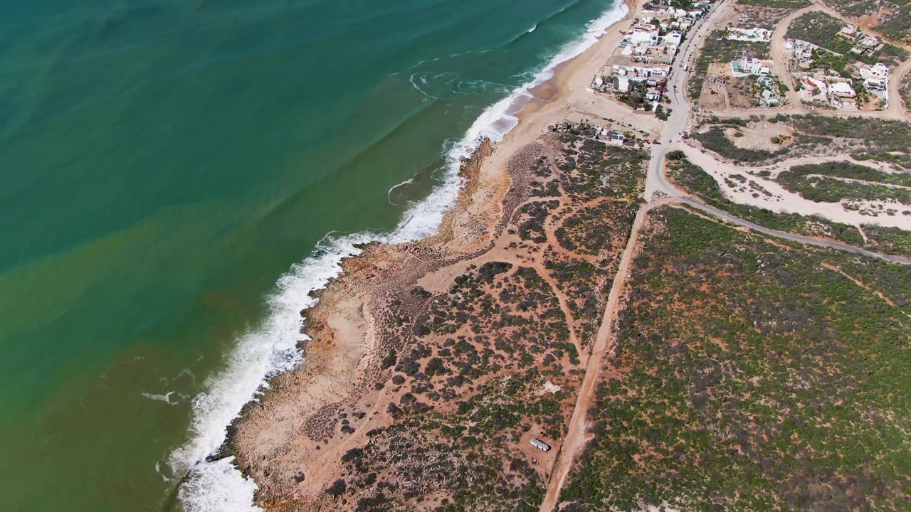 Aerial frame at Buzzards Beach East Cape BCS Mexico showing a wider stretch of green water, a white wave band, sandy and rocky shore, inland scrub, a dirt road, buildings, and no visible people.