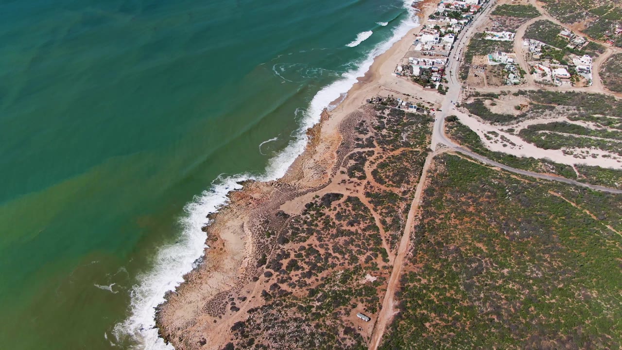 Aerial frame over Buzzards Beach, East Cape, BCS, Mexico, showing green ocean water, white surf, a sandy rocky shoreline, dirt roads, scrub vegetation, scattered buildings, and no visible people.