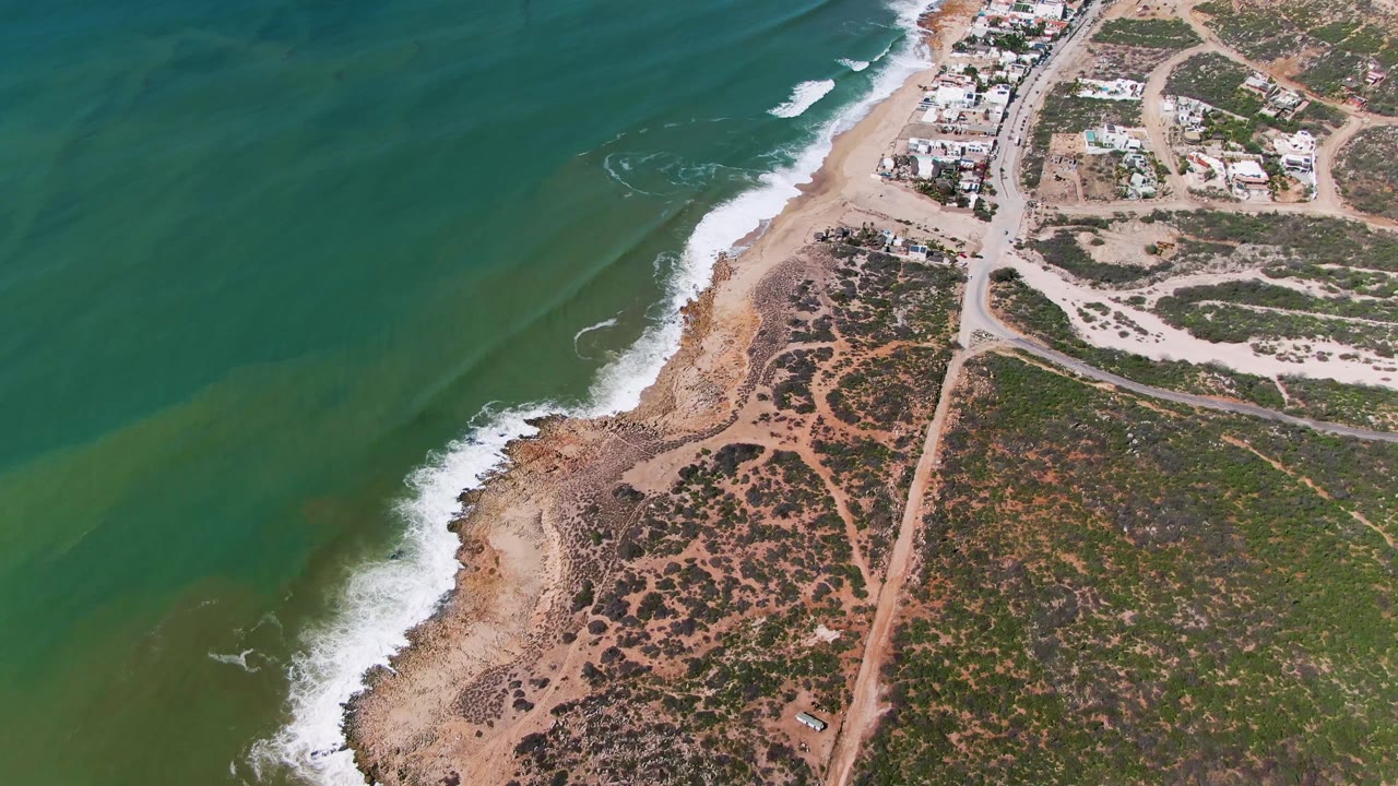 Drone view over Buzzards Beach, East Cape, BCS, Mexico, with waves curling toward the beach, a straight dirt road running inland, scrub-covered lots, scattered buildings, and no visible people.