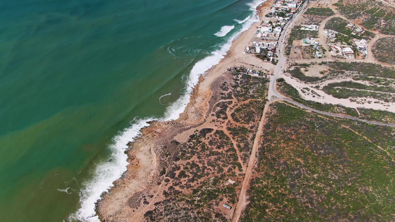 High aerial frame from Buzzards Beach, East Cape, BCS, Mexico, showing green water with a pale underwater band, white foam along the shore, road tracks, low vegetation, clustered buildings, and no visible people.