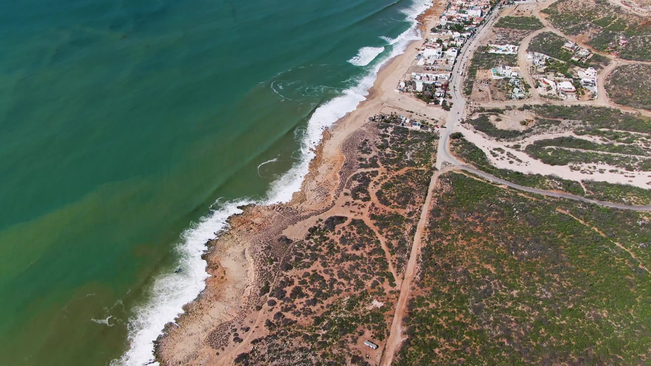 Aerial coastal scrub and dirt roads beside surf breaking on a narrow beach.