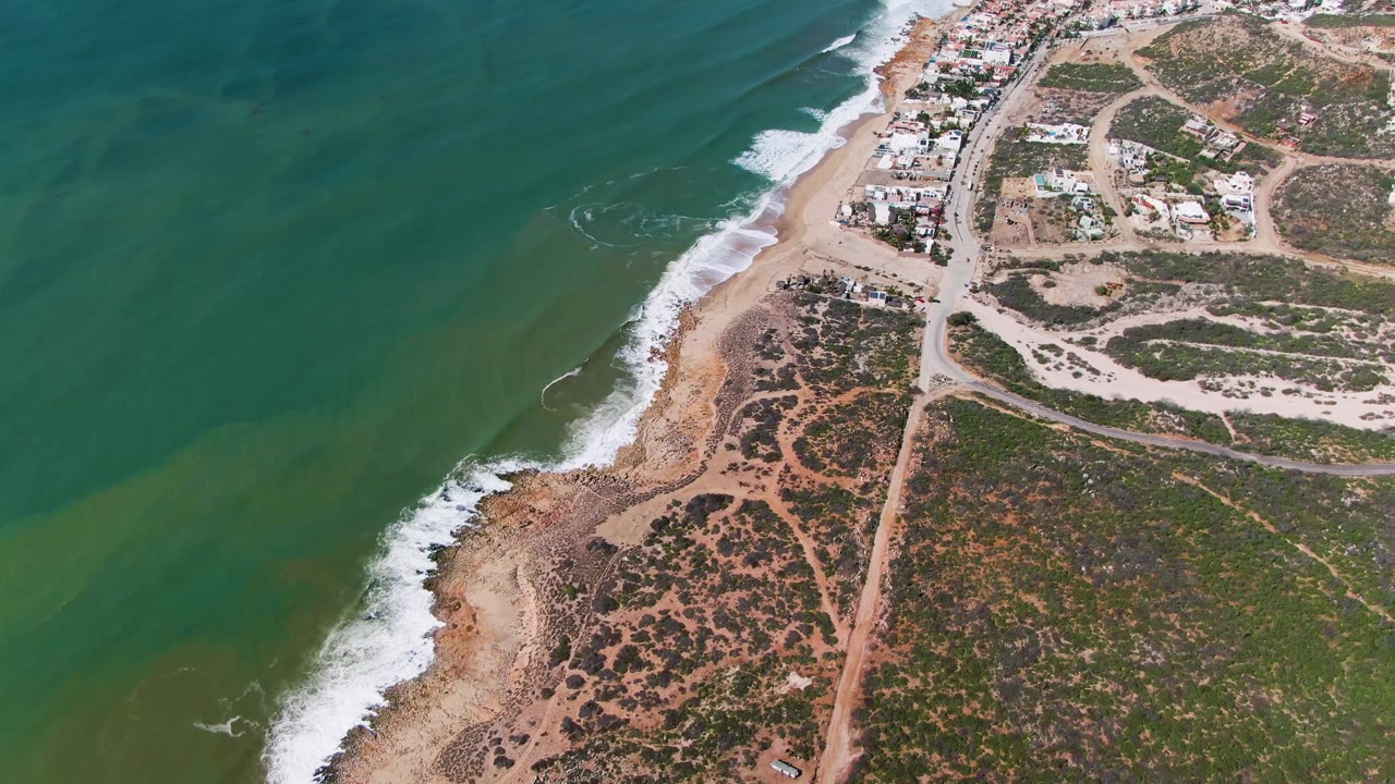 Opening aerial frame over Buzzards Beach East Cape BCS Mexico, showing green ocean water, white surf, sandy shore, scrub vegetation, dirt roads, small buildings, and no visible people.