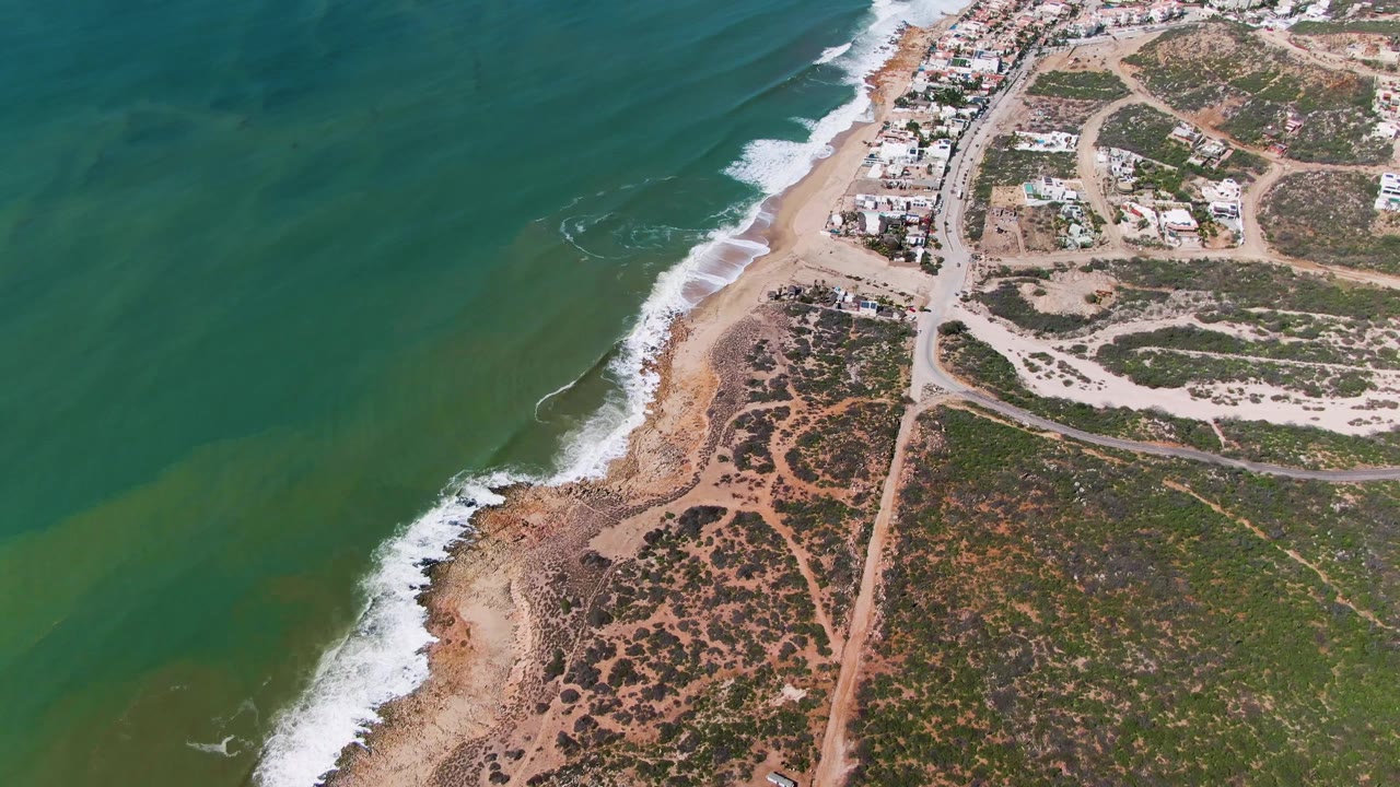 Drone view of surf curling along a sandy beach beside scrubland roads and coastal buildings