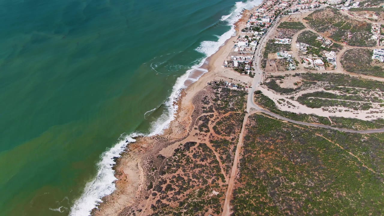 Aerial frame at 0:02 over Buzzards Beach East Cape BCS Mexico, showing the coast shifted slightly through the composition with foamy surf, green water, sandy beach, road cuts, low vegetation, buildings, and no visible people.