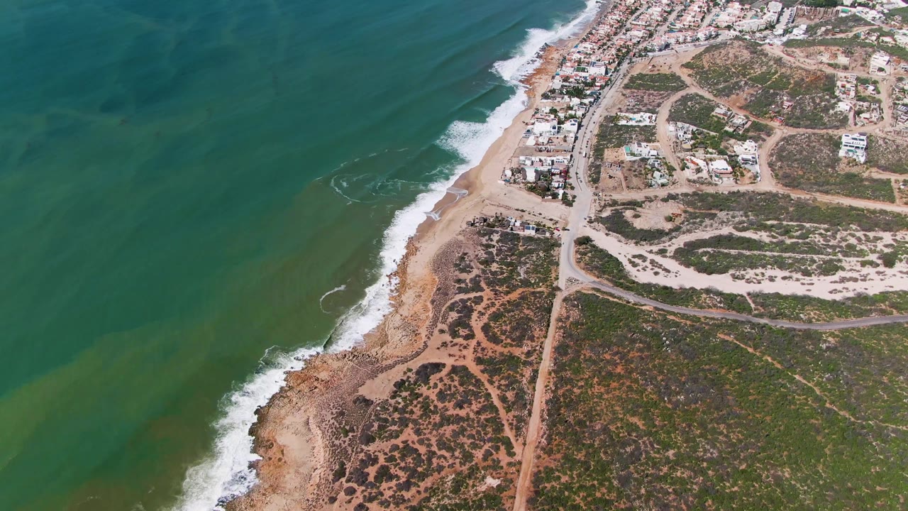 Aerial frame at Buzzards Beach East Cape BCS Mexico shows green ocean on the left, waves reaching a sandy and rocky beach, roads, scrubland, and residential buildings with no visible people.