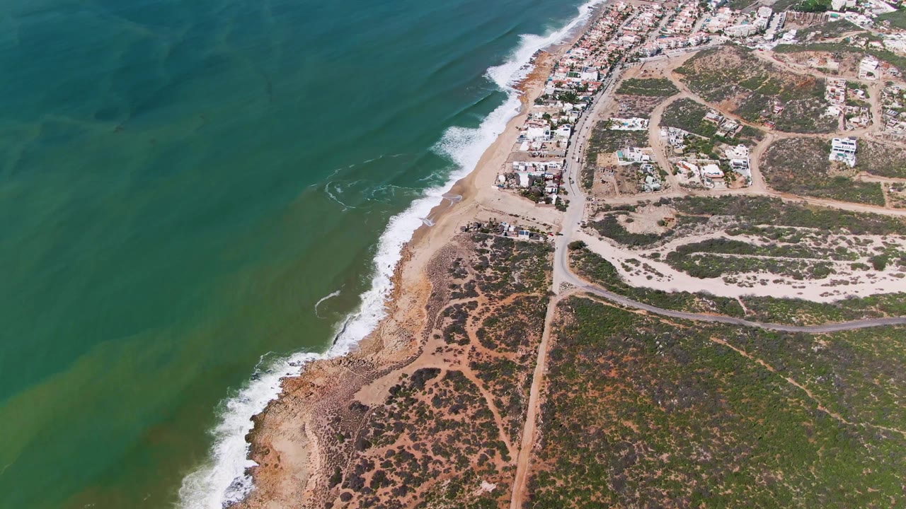 Aerial view at 0:01 shows the green water and breaking waves slightly farther along the beach, with dry vegetation, roads, and houses inland at Buzzards Beach East Cape BCS Mexico, with no visible people.