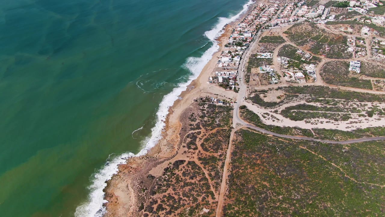 At 0:02 the aerial composition holds green ocean to the left and a curving surf edge through the center, with a beach road, dry lots, and residential buildings visible inland in Mexico, with no visible people.