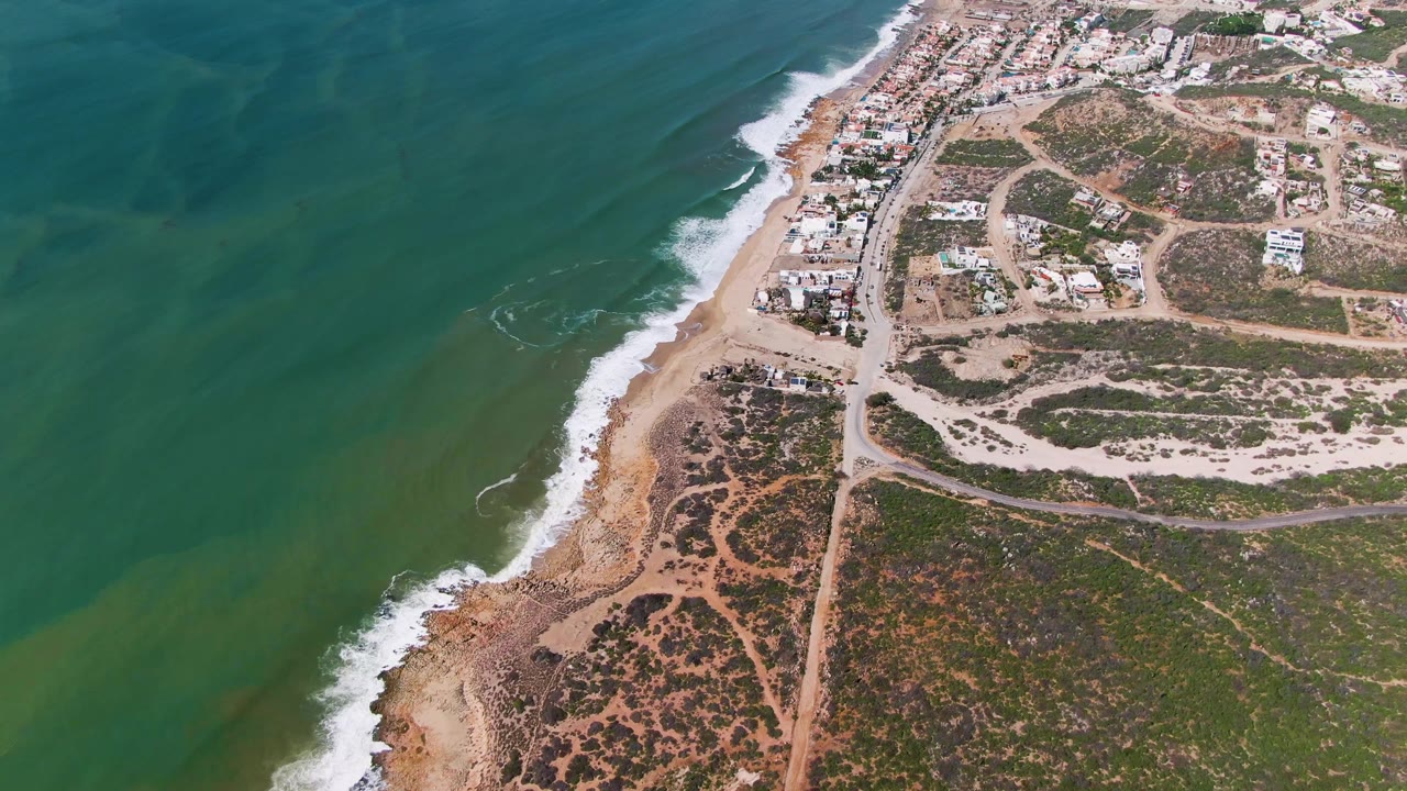 Aerial shoreline with green water, white surf, dirt tracks, brush, and coastal houses.