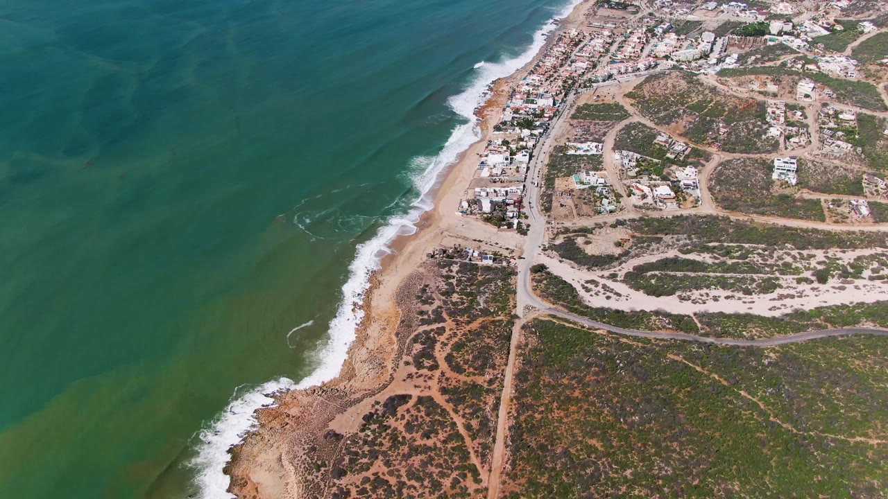 Opening aerial frame over Buzzards Beach East Cape BCS Mexico, with green-blue water on the left, surf along the sandy shoreline, roads and low buildings inland, and no visible people.