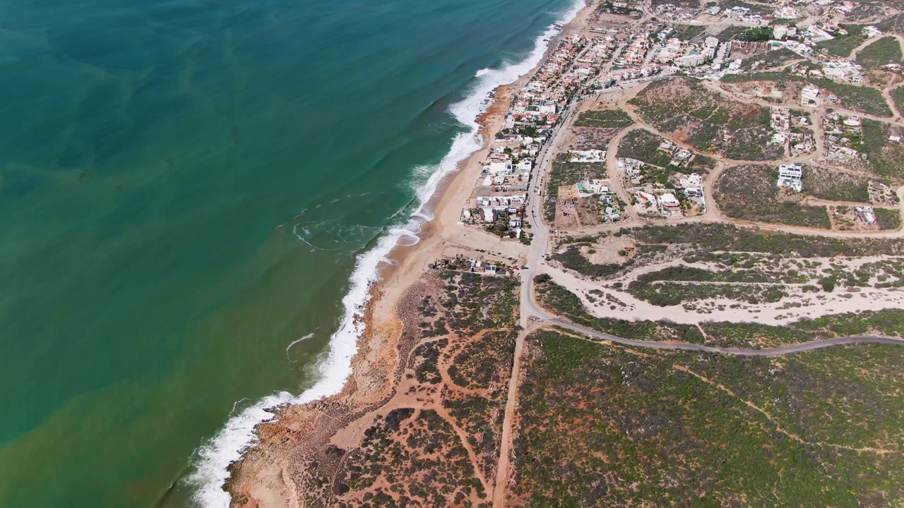 At 0:01, the Buzzards Beach shoreline remains centered as white water reaches the sand, with teal ocean to the left, dusty roads and scattered buildings to the right, and no visible people.