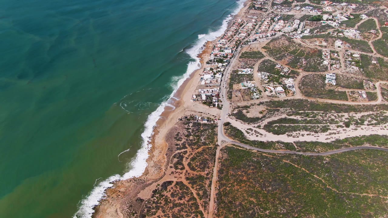Aerial coastline view with broad green water, beach break, road grid, and scrubland.