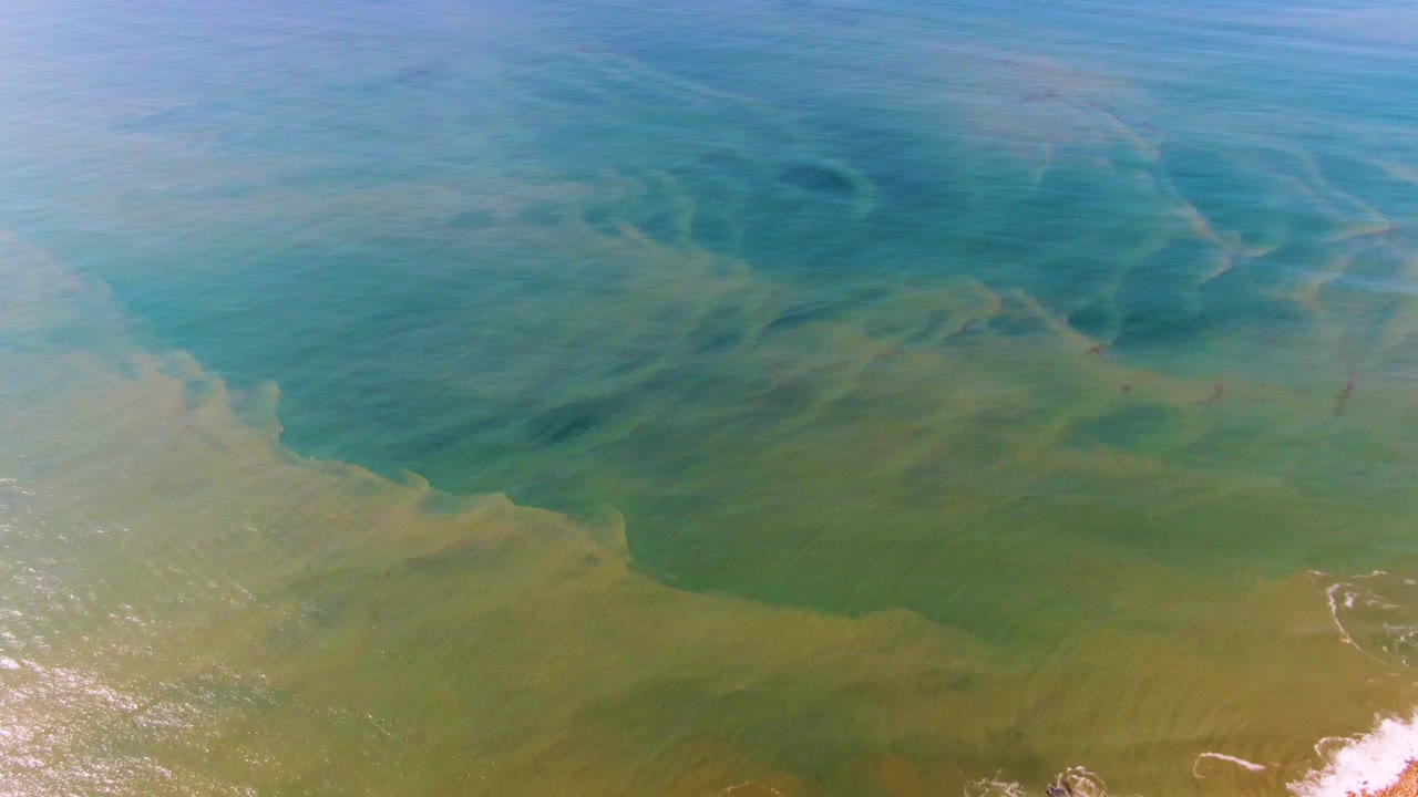 Overhead Buzzards Beach East Cape BCS Mexico water frame with blue-green bands, faint sandy plumes across the surface, and white foam entering along the lower right edge, no visible people.