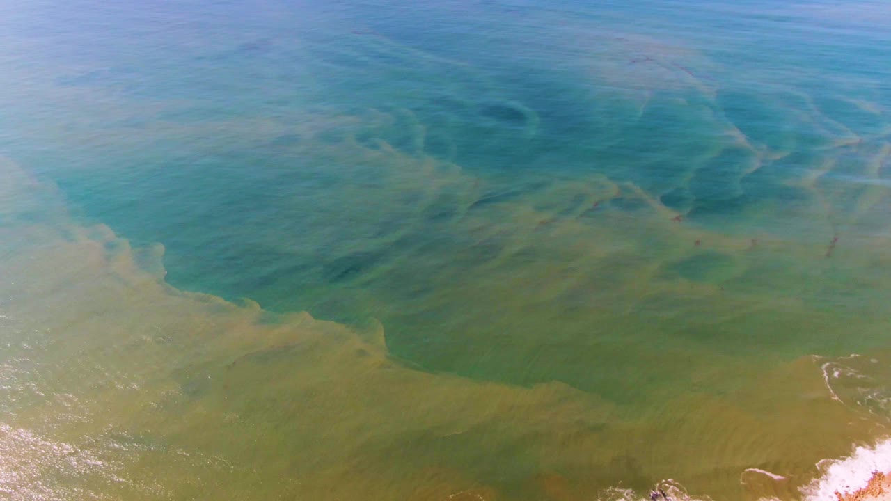 Aerial view over Buzzards Beach East Cape BCS Mexico showing turquoise water, darker blue patches, soft green shallows, and a small white breaking wave at the lower right, no visible people.