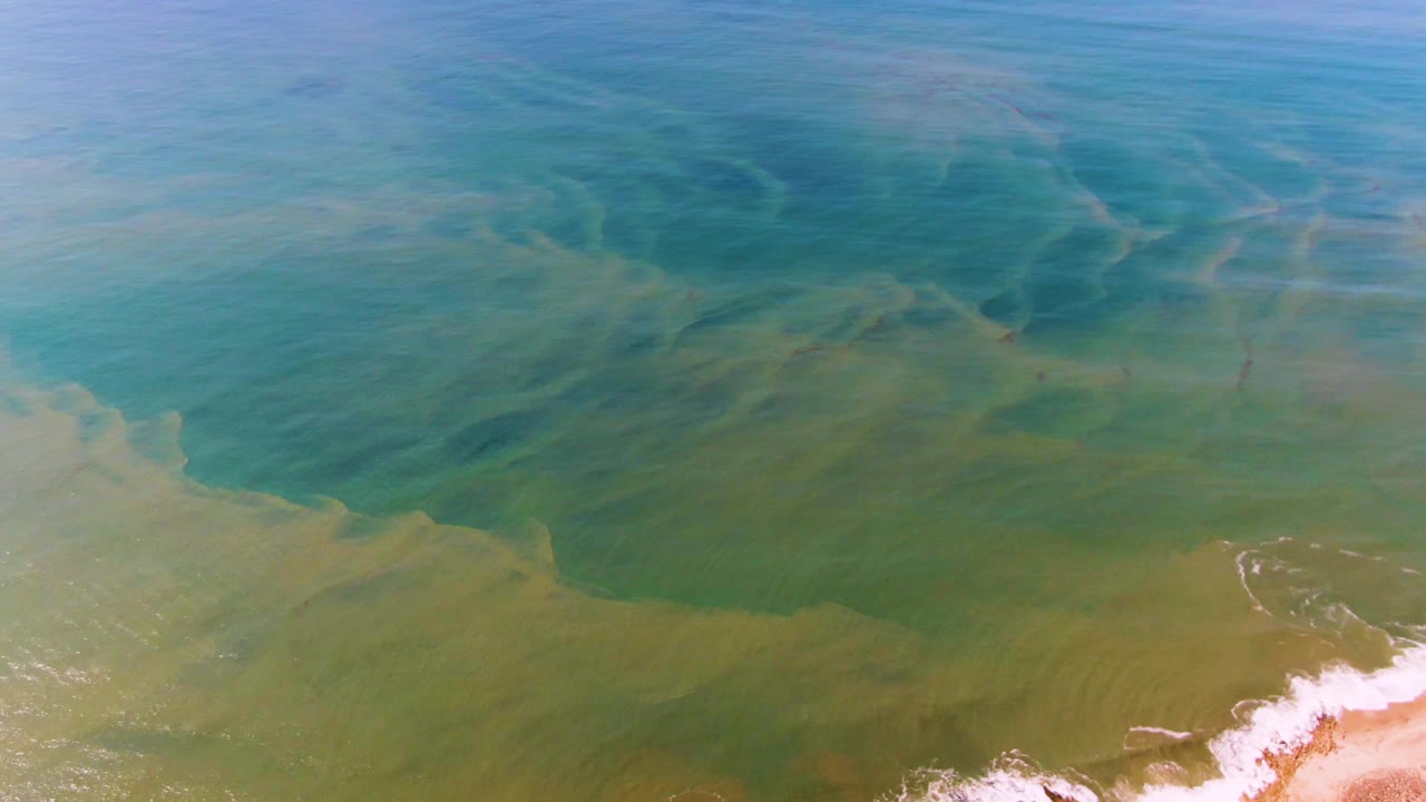 Aerial frame at Buzzards Beach East Cape BCS Mexico showing shallow blue-green water, visible seabed patterns, a sandy lower edge, and no visible people.