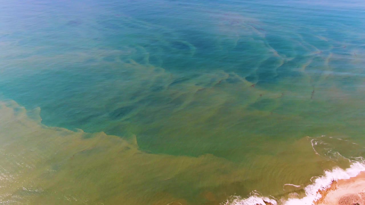 Second aerial frame over Buzzards Beach East Cape BCS Mexico with darker submerged seabed bands, pale green shallows, white foam at the beach edge, and no visible people.