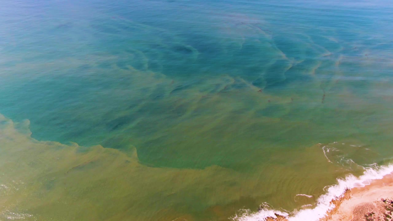 Mid-clip aerial frame from Buzzards Beach East Cape BCS Mexico showing broad blue-green water, darker seabed streaks, low white foam near sand, and no visible people.