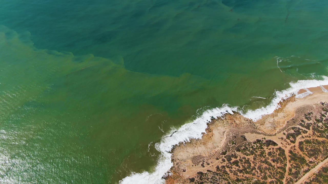 Green water and white foam meet a rocky sandy shoreline.