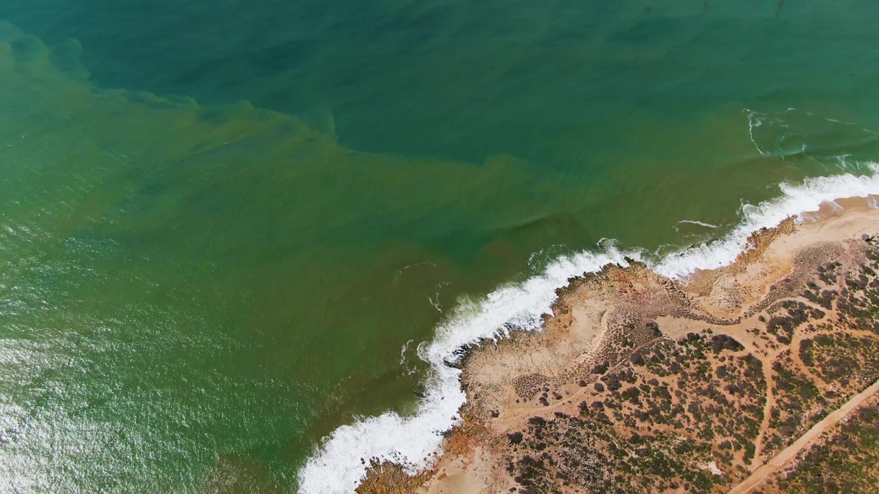 Aerial coast view with darker green water, foam line, sandy point, scrub, and dirt track.