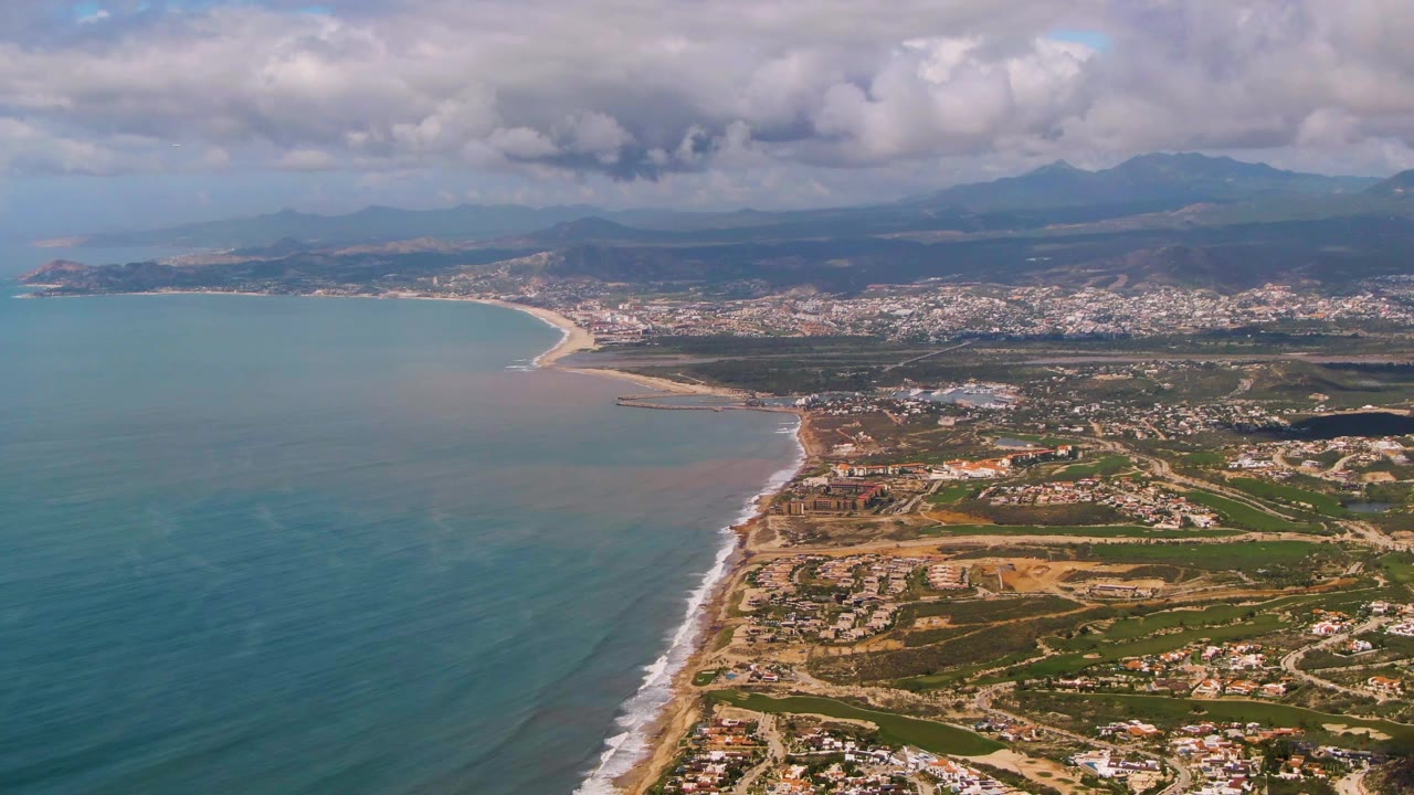 Aerial view over Buzzards Beach East Cape BCS Mexico with turquoise ocean at left, a sandy shoreline, inland buildings, distant mountains, layered clouds, and no visible people.