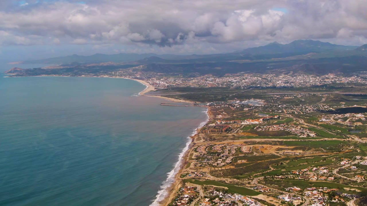 Drone view above Buzzards Beach East Cape BCS Mexico showing blue-green water, a narrow surf line, coastal development, hills and mountains under broken cloud cover, and no visible people.