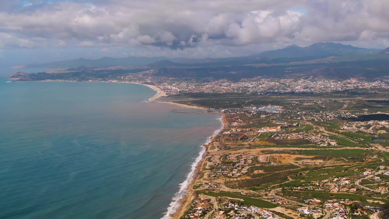 Wide aerial frame from Buzzards Beach East Cape BCS Mexico with open ocean on the left, the beach curving toward a coastal town, mountains in the distance, heavy clouds, and no visible people.