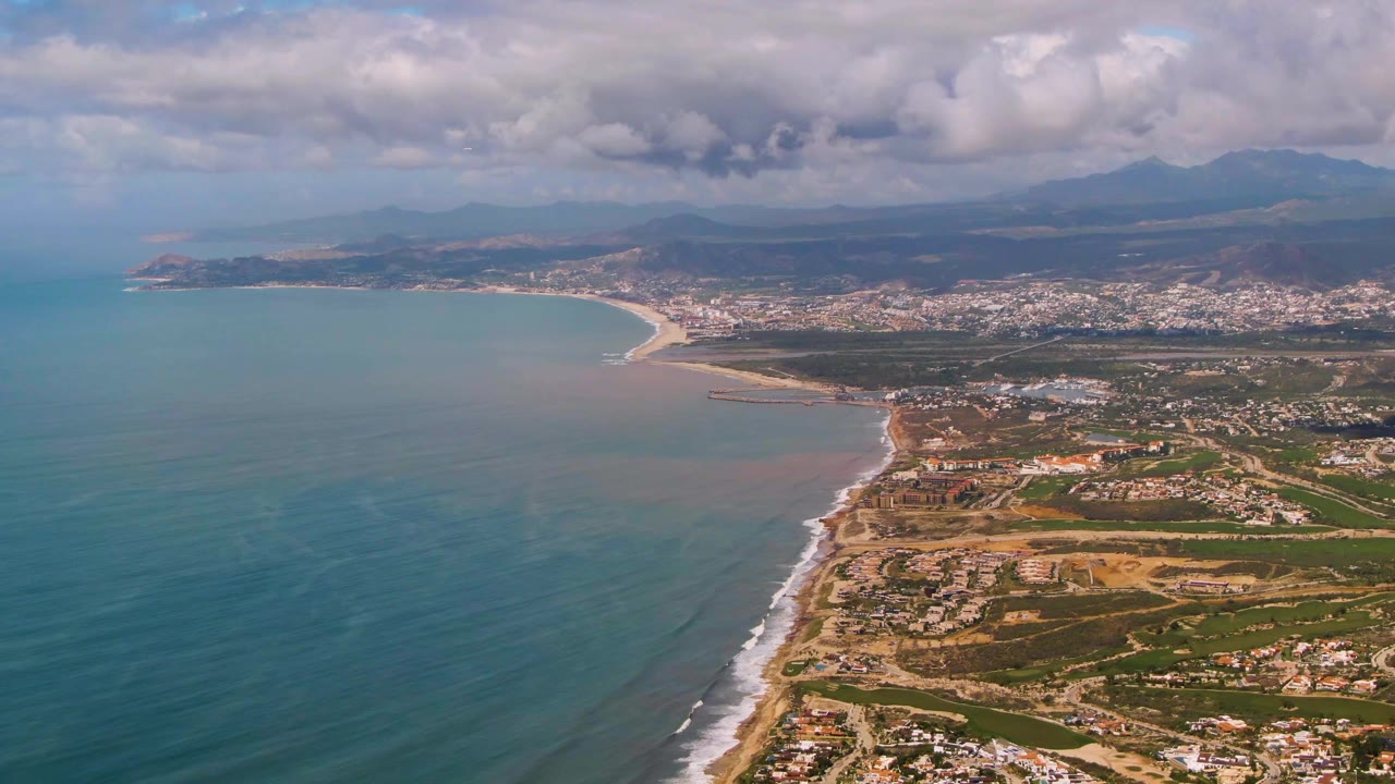 Aerial view over Buzzards Beach East Cape BCS Mexico with blue-green ocean at left, a sandy shoreline, coastal development inland, distant mountains, layered clouds, and no visible people.