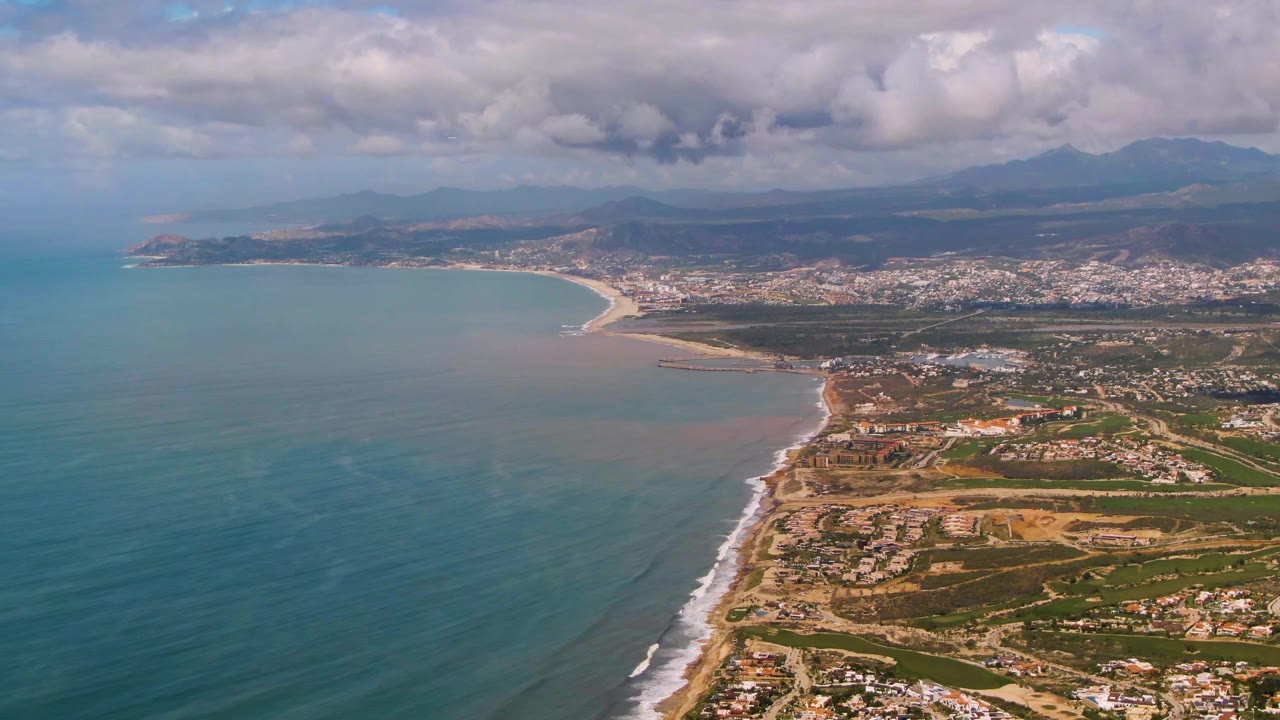 High aerial frame of Buzzards Beach East Cape BCS Mexico showing the beach curve more centrally, pale surf along the shore, urban development beyond it, mountains on the horizon, and no visible people.