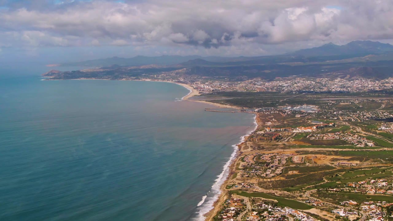 Drone-height view over Buzzards Beach East Cape BCS Mexico with open water filling the left side, surf tracing the shoreline, a coastal city in the background, hazy mountains, broken cloud cover, and no visible people.