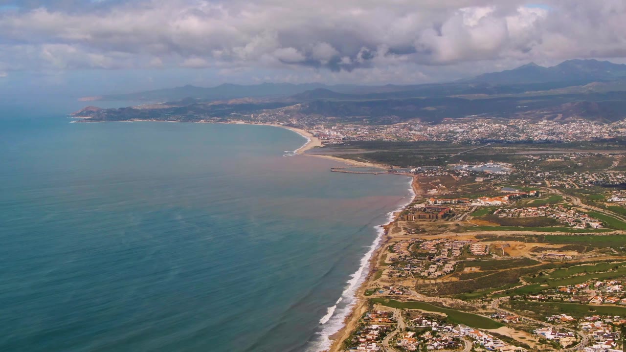 Aerial coastal landscape with teal water, surf line, beach, city, mountains, and clouds.