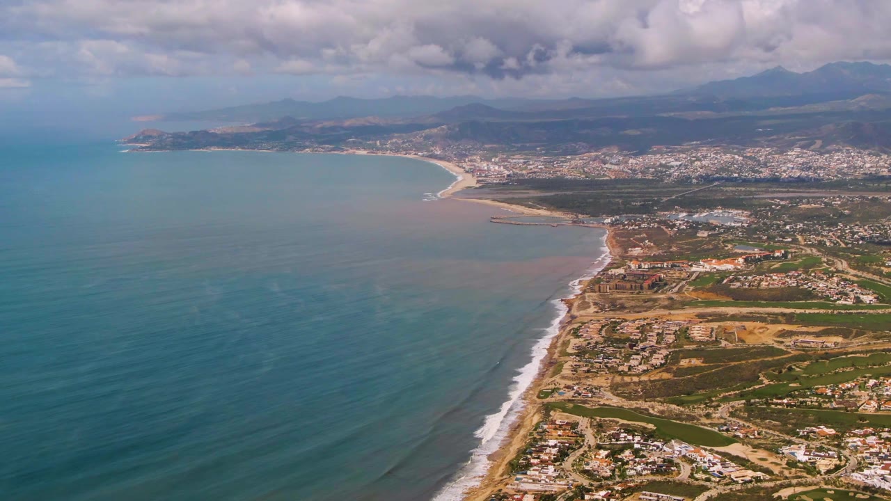 Opening aerial frame over Buzzards Beach East Cape BCS Mexico, with blue-green ocean at left, surf along the sandy coast, town and mountains in the distance, and no visible people.