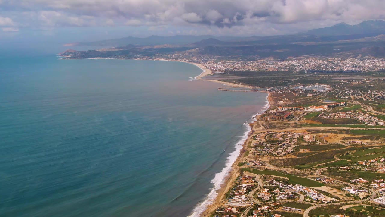 Mid-clip aerial frame of Buzzards Beach East Cape BCS Mexico, with the bay curving toward a distant town, a bright surf edge on the beach, clouded mountains beyond, and no visible people.
