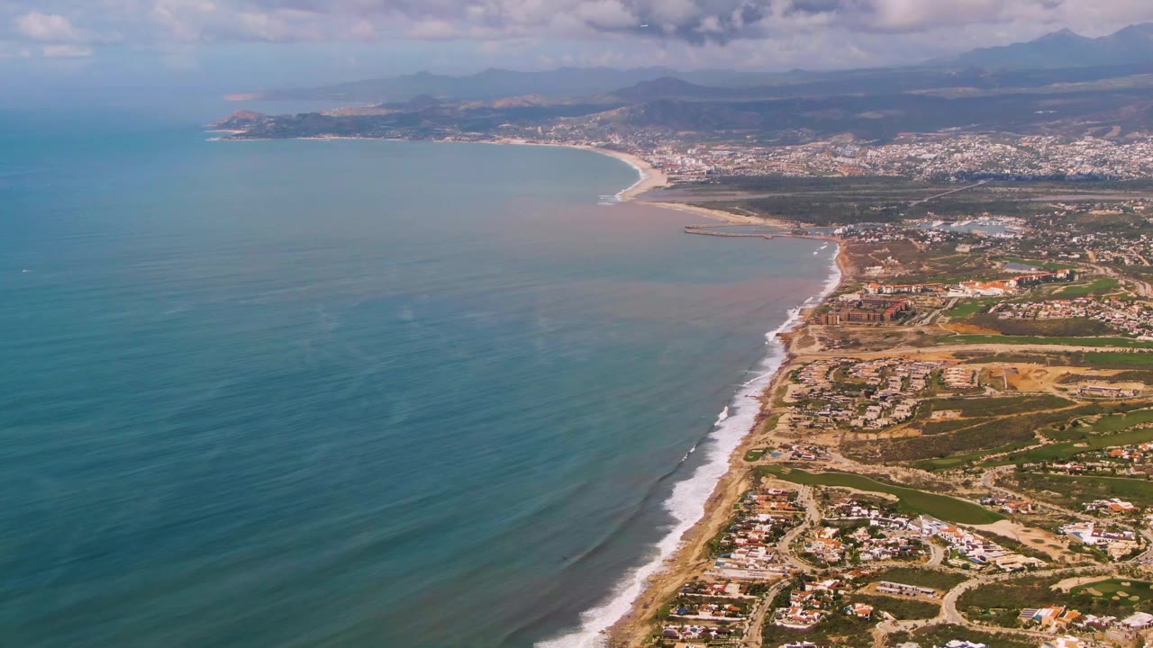 Opening aerial frame over Buzzards Beach East Cape BCS Mexico, with blue-green ocean on the left, white surf along the sand, coastal housing on the right, distant town and mountains, and no visible people.