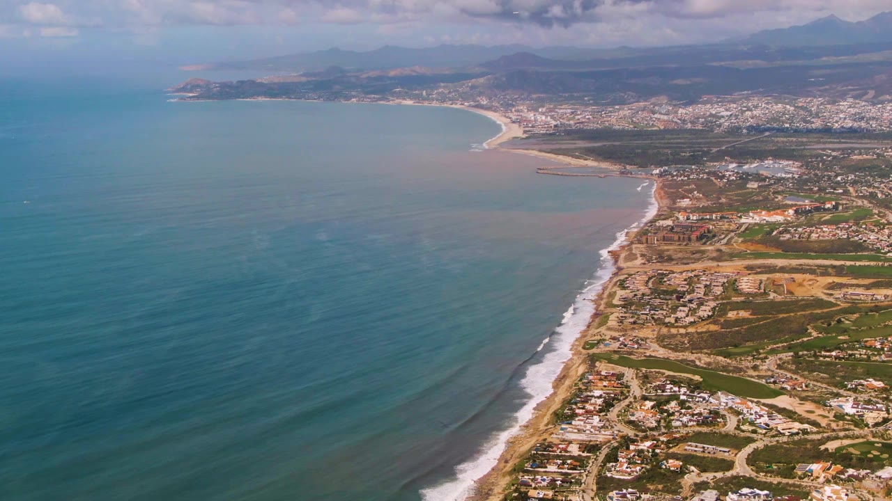 Aerial frame over the Buzzards Beach shoreline in Mexico, showing the diagonal surf line, nearshore water color changes, houses and roads along the coast, distant town blocks, mountains, and no visible people.