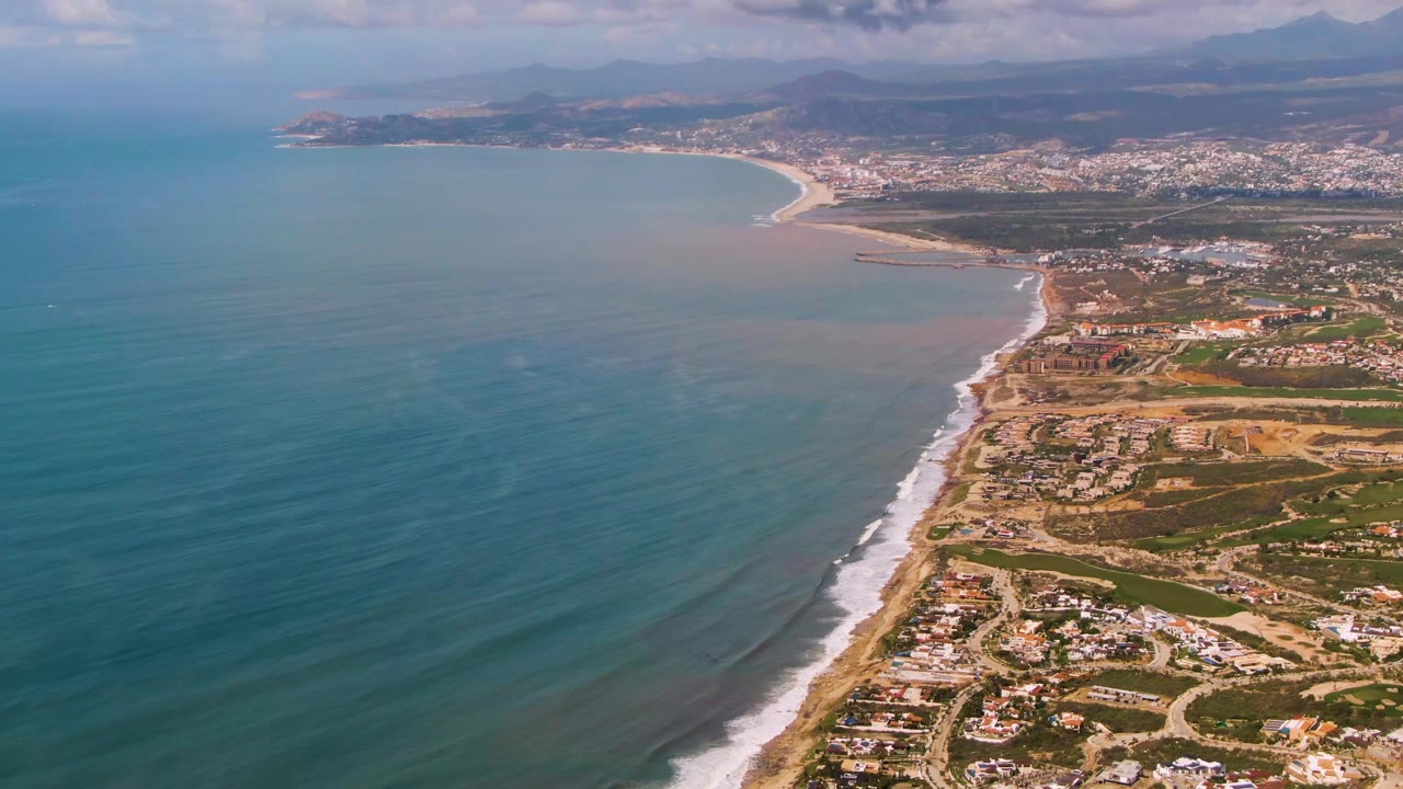 Mid-clip aerial frame at Buzzards Beach East Cape BCS Mexico, with broad ocean texture, a narrow white wave band on the sand, residential development along the right edge, a distant coastal town, hazy mountains, and no visible people.
