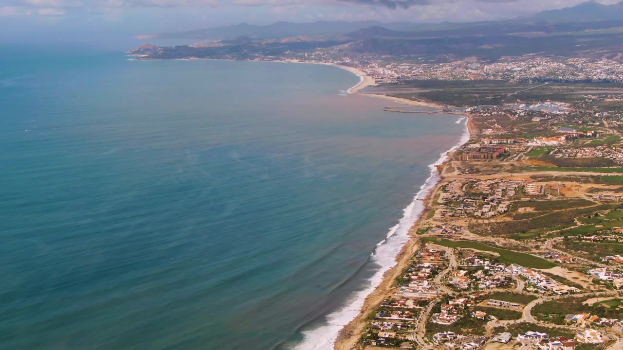 Aerial coastline frame with wide ocean, breaking surf, beach homes, town, and mountains.