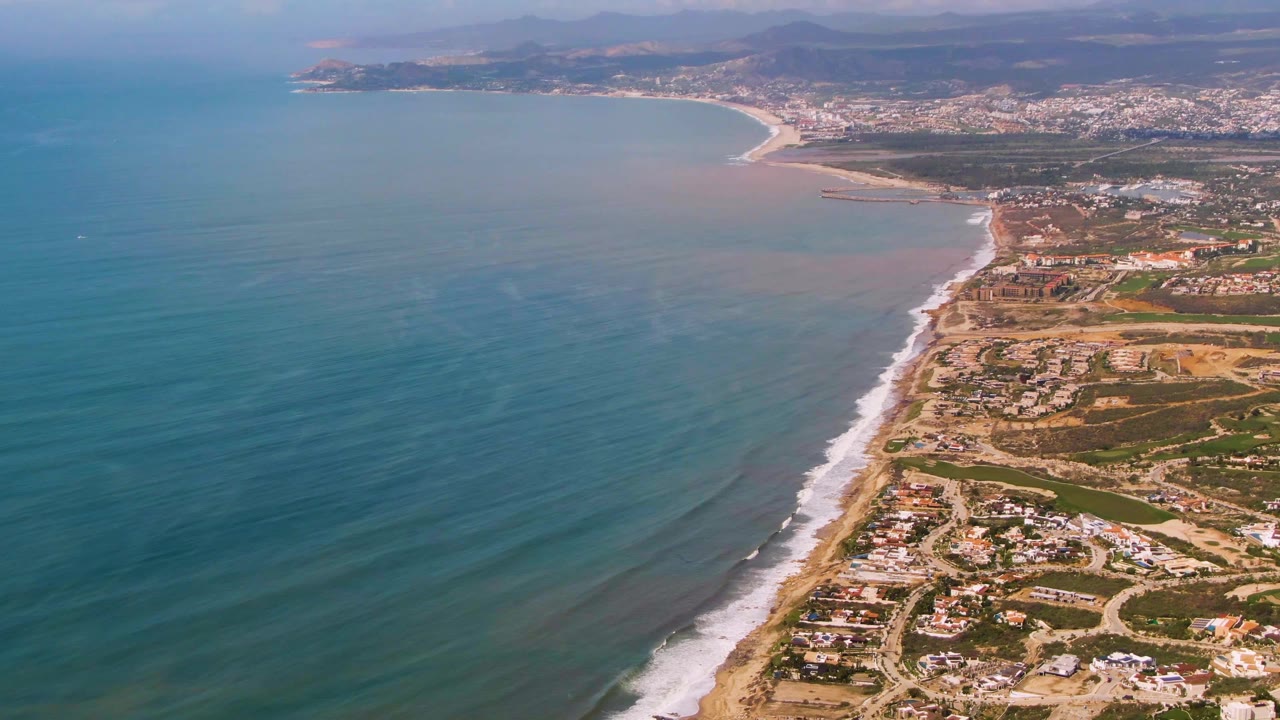 Aerial coastline with blue-green ocean, sandy beach, surf, roads, and coastal buildings.