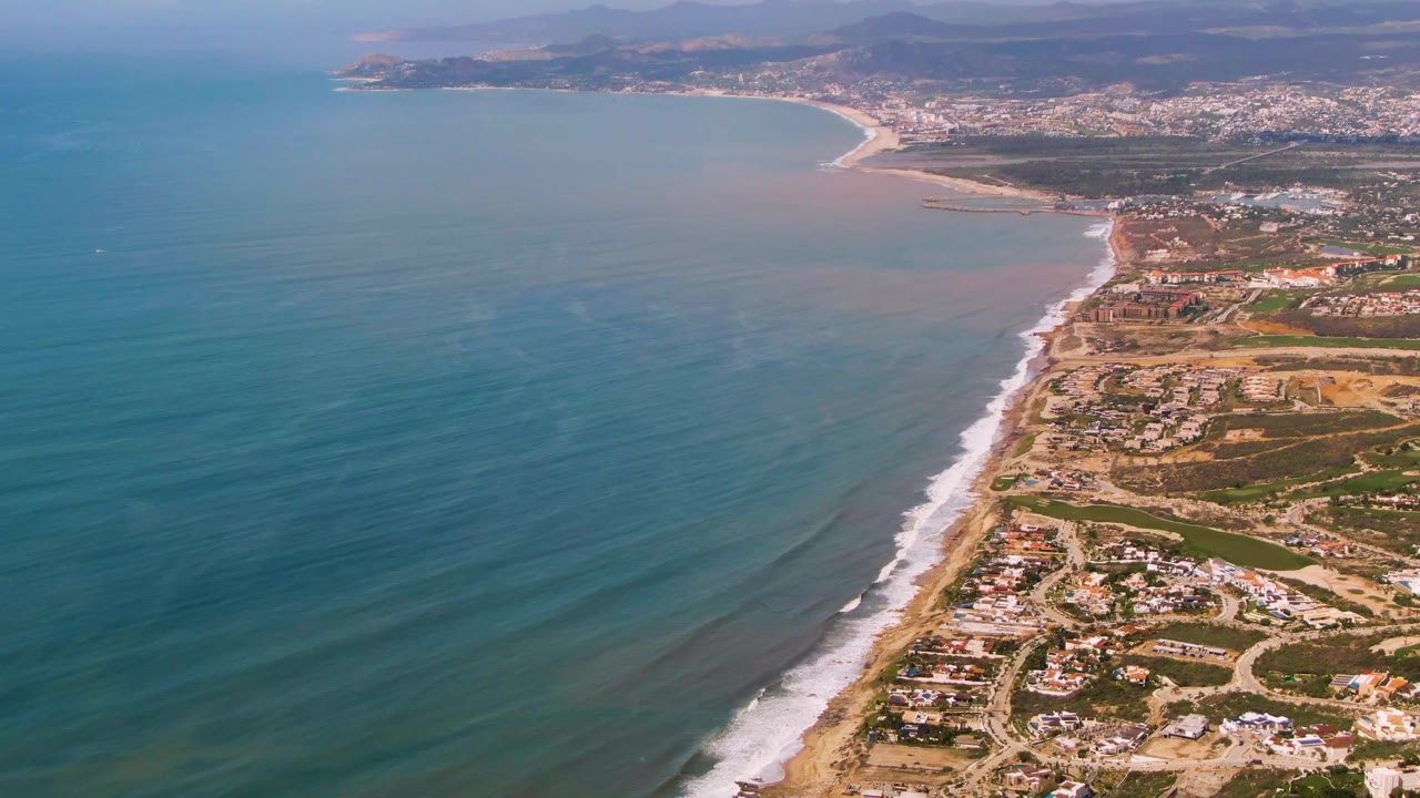Drone view over Buzzards Beach East Cape BCS Mexico showing the shoreline running diagonally through frame, calm ocean water, white wave foam, beach roads, buildings, and no visible people.