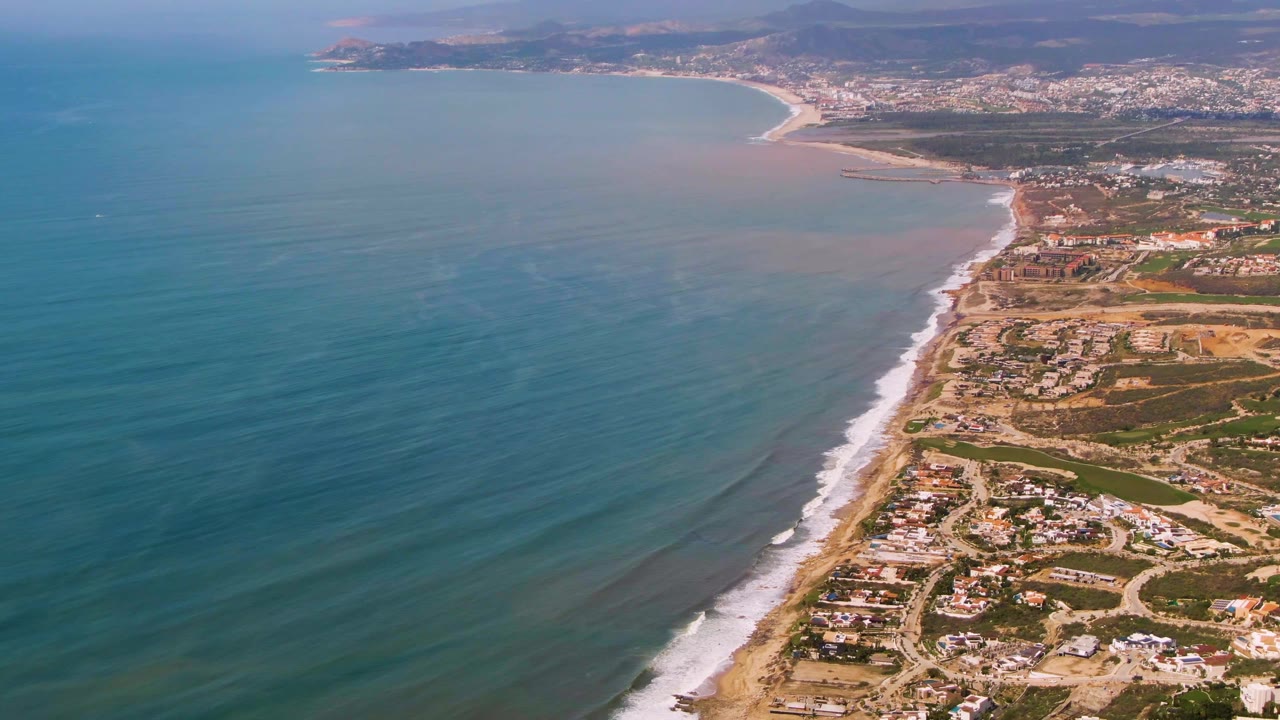 High aerial frame from Buzzards Beach East Cape BCS Mexico with a curved bay and town in the distance, turquoise water to the left, surf along the beach, developed lots to the right, and no visible people.