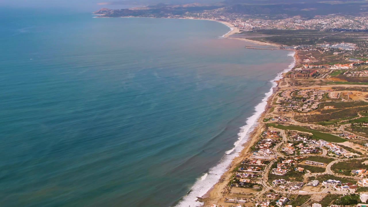 Aerial view of Buzzards Beach East Cape BCS Mexico with textured ocean swells on the left, a bright surf edge along the sand, scattered coastal buildings and roads on the right, and no visible people.