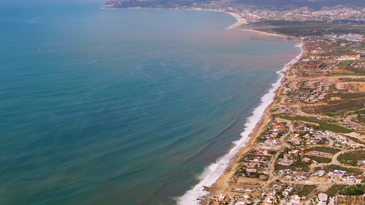 High aerial view over Buzzards Beach East Cape BCS Mexico with blue ocean on the left, a sandy surf line, coastal homes and roads on the right, and no visible people.