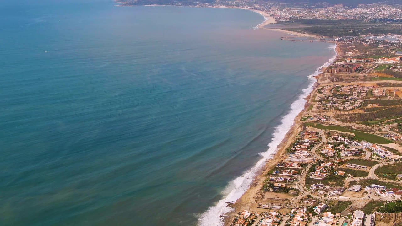 Aerial frame over Buzzards Beach East Cape BCS Mexico showing bands of blue water, white waves meeting the sand, gridded roads, scattered coastal buildings, and no visible people.
