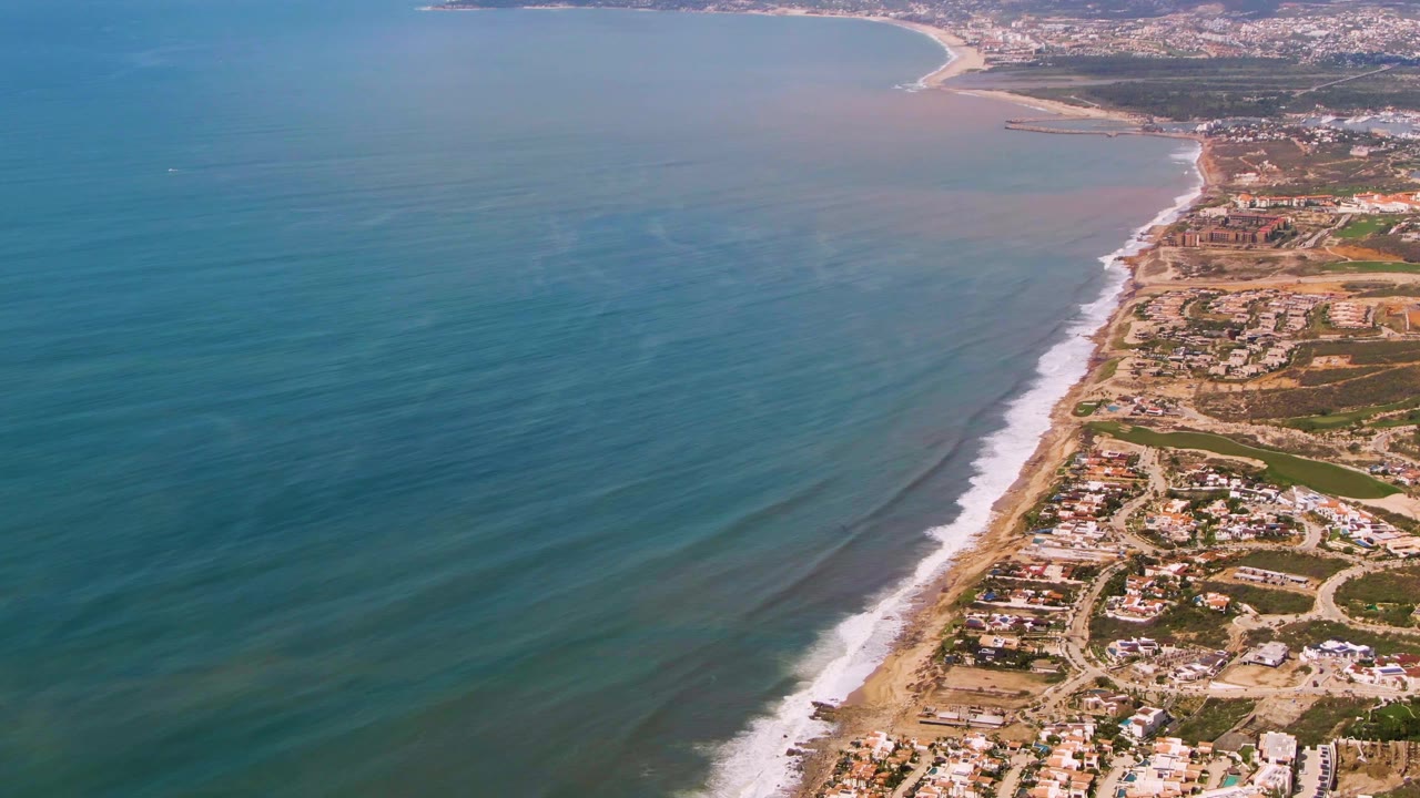 Wide drone view of Buzzards Beach East Cape BCS Mexico with turquoise-blue ocean taking most of the frame, a narrow sandy beach, coastal houses, roads, and no visible people.